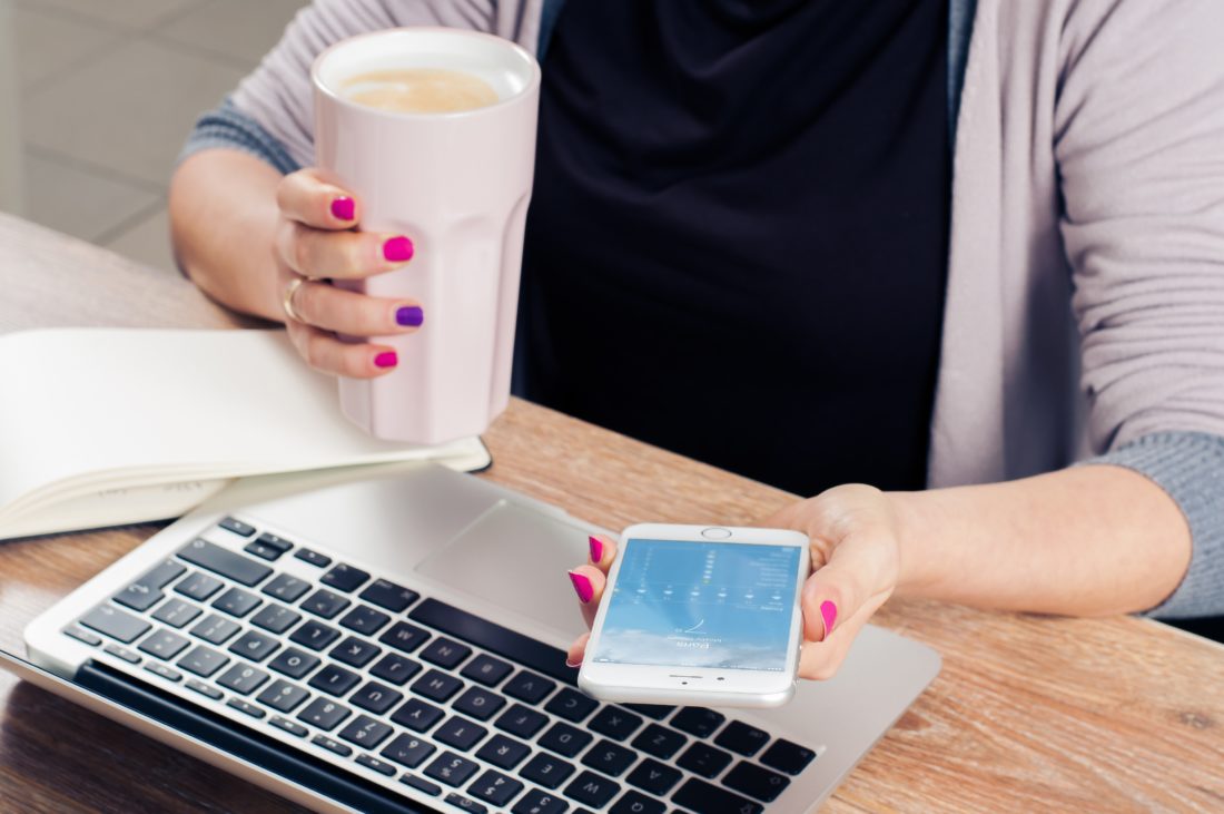 Woman holding a coffee in her right hand and an iPhone in her left hand with a laptop computer on the desk in front of her